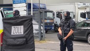 Zollbeamte stehen mit Schild und Waffe vor einem Firmengelände. © NDR 