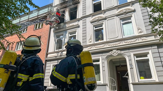 Zwei Feuerwehrleute stehen vor dem Wohnhaus in der Hopfenstraße, in dem am Mittag ein Feuer ausgebrochen war. © NDR Foto: Elias Bartl