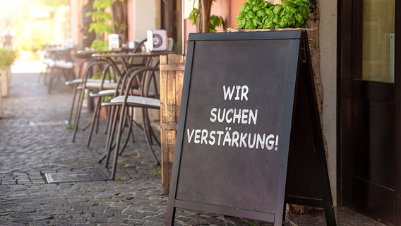 Vor einem Restaurant steht ein Schild mit der Aufschrift "Wir suchen Verstärkung!". © picture alliance / CHROMORANGE / Foto: Michael Bihlmayer