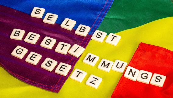 Buchstabenwürfel zeigen das Wort Selbstbestimmungsgesetz auf einer Regenbogenfahne. © picture alliance / CHROMORANGE Foto: Udo Herrmann
