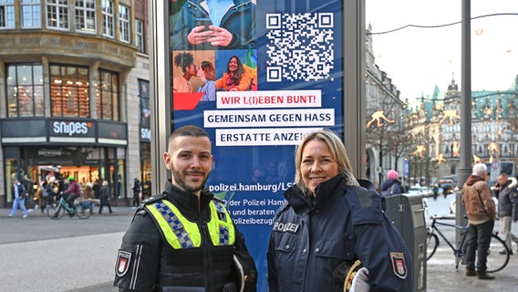 Tobias Conrad (l) und Miria Lottmann, die LSBTI*-Ansprechpersonen der Polizei Hamburg, stehen vor einer digitalen Litfaßsäule mit einem Motiv der Kampagne "Wir l(i)eben bunt". © Niklas Graeber/dpa Foto: Niklas Graeber/dpa