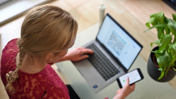 Eine Frau sitzt zu Hause an einem Glastisch und tippt auf einem Laptop, in der rechten Hand hält sie ein Smartphone. © picture alliance / dpa Foto: Helena Dolderer