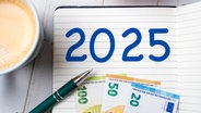 In einem Block steht die Jahreszahl "2025" daneben liegen ein Stift und Geldscheine. Links am Bildrand steht eine Kaffeetasse. © picture alliance / CHROMORANGE 