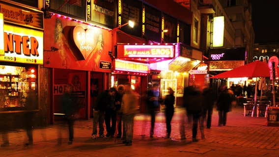 Eine Aufnahme vom Hamburger Kiez bei Nacht. Im Hintergrund sind die Lokalitäten zu sehen, davor gehen einige Menschen zu Fuß, die nicht zu erkennen sind. © imageBROKER Foto: Stefan Huwiler