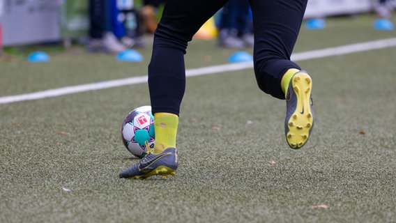 Auf einem Kunstrasenplatz liegt ein Fußball, daneben steht ein Spieler mit Fußballschuhen. © picture alliance / Fotostand 