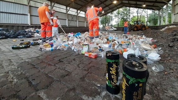 Mitarbeiter der Stadtreinigung sortieren in einer Halle Lachgasflaschen aus einem Müllhaufen heraus. Mehrere Lachgasflaschen stehen im Vordergrund. © Stadtreinigung Hamburg 