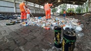 Mitarbeiter der Stadtreinigung sortieren in einer Halle Lachgasflaschen aus einem Müllhaufen heraus. Mehrere Lachgasflaschen stehen im Vordergrund. © Stadtreinigung Hamburg 