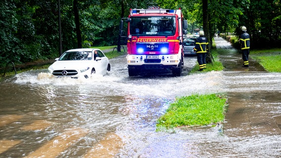 Ein Auto fährt durch eine überflutete Straße in Hamburg-Lohbrügge, in der ein Feuerwehrwagen parkt. © dpa Foto: Daniel Bockwoldt