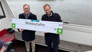 Robert Henrich, Vorstandsvorsitzender Hochbahn und Hamburgs Verkehrssenator Anjes Tjarks (Grüne) halten das Schild für die neue U4-Haltestelle "Moldauhafen" hoch. © NDR Foto: Anna Rüter