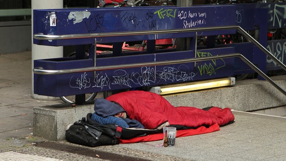 Ein Obdachloser liegt mit Schlafsack an der S-Bahn Reeperbahn auf dem Bürgersteig und schläft. © IMAGO Foto: Hanno Bode