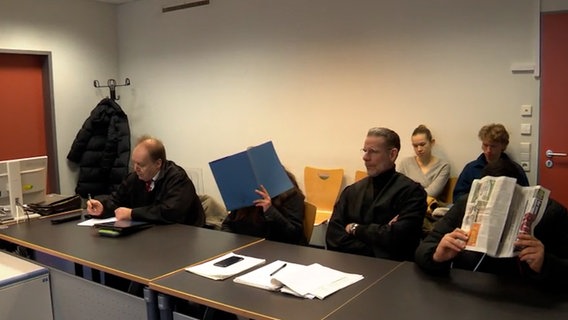 Angeklagte sitzen mit Mappe und Zeitung vor dem Gesicht im Gerichtssaal. © NDR 