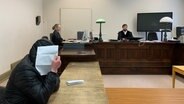 Ein Mann bedeckt in einem Gerichtssaal sein Gesicht mit einem Blatt Papier. © NDR / Elke Spanner 