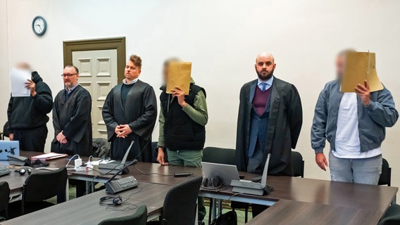 Vor dem Landgericht Hamburg beginnt ein Prozess gegen drei Männer wegen versuchter Freiheitsberaubung und wegen Drogenhandels. Die Angeklagten stehen neben ihren Anwälten und verdecken ihre Gesichter. © picture alliance / dpa Foto: Jonas Walzberg