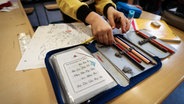 Ein Schüler greift nach einem Stift aus seiner Stiftemappe mit einem Zettel mit den Buchstaben des Alphabets in einer 2. Klasse in der Grundschule Hasselbrook. © picture alliance/dpa | Christian Charisius Foto: Christian Charisius