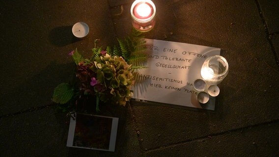 Ein Schild mit der Aufschrift "Für eine offene und tolerante Gesellschaft - Antisemitismus hat hier keinen Platz" liegt neben Kerzen und Blumen auf dem Gehweg nahe der Synagoge.  Foto: Jonas Walzberg