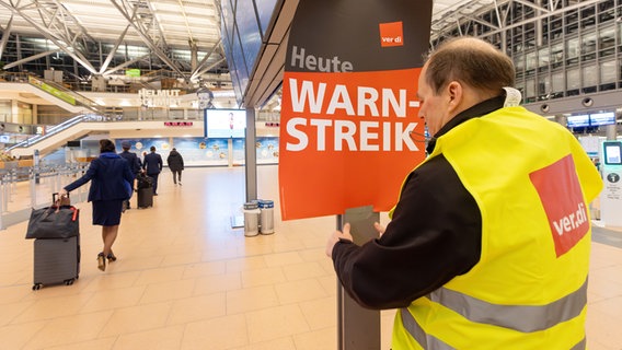 Ein Vertreter der Gewerkschaft ver.di hängt ein Plakat mit der Aufschrift "Warnstreik" in einem Terminal im Hamburger Flughafen auf. © picture alliance/dpa Foto: Bodo Marks