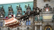 Eine Person in einem Weihnachtsmannkostüm winkt auf einem Schlitten mit Rentieren, der an einem Seil über den Weihnachtsmarkt hängt, in die Menge vor dem Hamburger Rathaus. © picture alliance / dpa Foto: Niklas Graeber