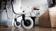 Ein Rollator steht in einer Wohnung, im Hintergrund ist ein älterer Mann zu erkennen. © IMAGO / photothek 