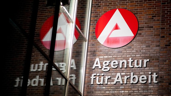 Eine Backsteinfassade mit Logo und Schriftzug der Agentur für Arbeit spiegelt sich in einer Glasfront. © Hauke-Christian Dittrich/dpa Foto: Hauke-Christian Dittrich/dpa