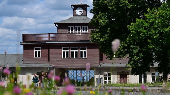 Ein Eingangstor mit einem kleinen Turm, auf dem eine Uhr abgebildet ist. Im Vordergrund stehen Bäume und wachsen Blumen. © picture alliance/dpa | Martin Schutt Foto: Martin Schutt