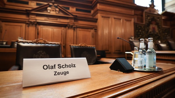 Ein Schild mit dem Text "Olaf Scholz Zeuge" steht auf einem Platz auf der Senatsbank vor Beginn einer Sitzung des Parlamentarischen Untersuchungsausschusses "Cum-Ex" im Plenarsaal der Hamburgischen Bürgerschaft im Rathaus. © picture alliance/dpa Foto: Christian Charisius