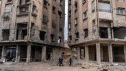 Menschen gehen zwischen Gebäuden, die durch Luftangriffe während der Assad-Herrschaft in Douma am Stadtrand von Damaskus beschädigt wurden. © Mosa'ab Elshamy/AP/dpa 