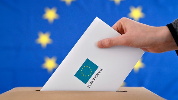 Ein Wahlbrief wird in eine Wahlurne geworfen, im Hintergrund ist eine EU-Flagge zu sehen.  © picture Alliance / Panama Pictures 