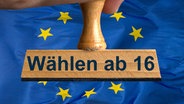 Ein symbolischer Holzstempel mit der Aufschrift "Wählen ab 16", wird von einer Hand im Anschnitt vor einer EU-Flagge gehalten. © picture alliance / SULUPRESS.DE | Torsten Sukrow / SULUPRESS.DE 