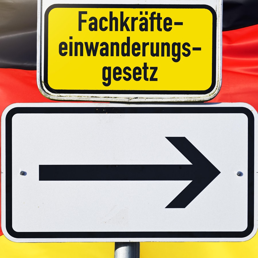 Ein Schild mit einem Pfeil und der Aufschrift Fachkräfteeinwanderungsgesetz vor einer deutschen Flagge © picture alliance / CHROMORANGE Foto: Christian Ohde