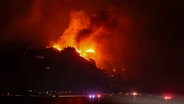 Flammen eines Großbrandes in Los Angeles sind aus einiger Entfernung im Dunklen auf einem Hügel zu sehen. © dpa bildfunk/AP Foto: Etienne Laurent