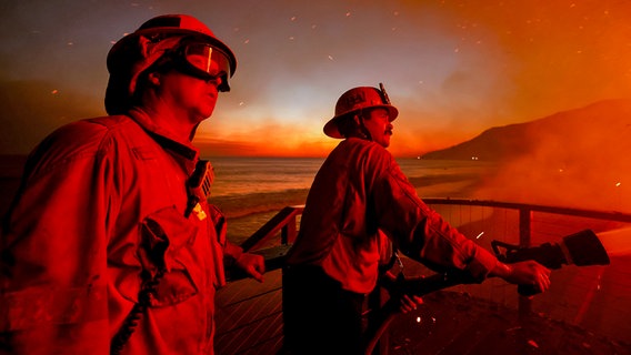 Feuerwehrleute stehen vor einem brennenden Haus, rundherum sind Flammen und Qualm zu sehen. © dpa bildfunk/AP Foto: Etienne Laurent