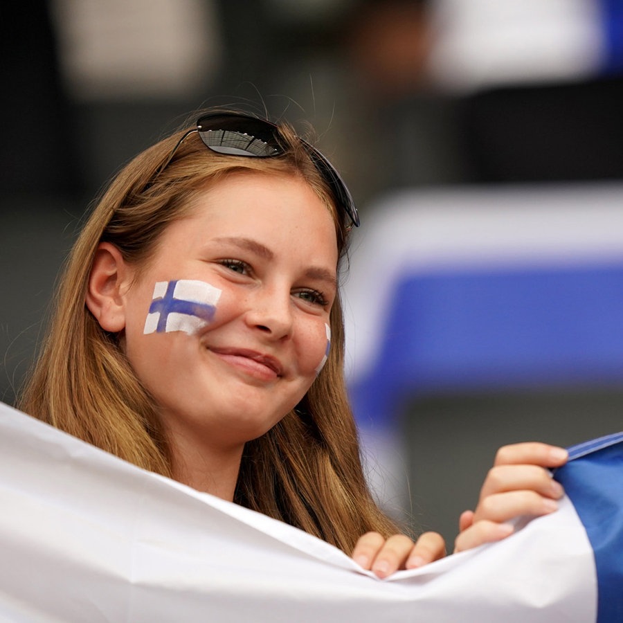 Zwei Mädchen halten lächelnd eine finnische Flagge hoch. Auf ihren Wangen sind ebenfalls finnische Flaggen gemalt. © PA Wire/dpa Foto: Tim Goode