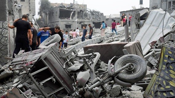 Mehrere Menschen stehen vor Trümmern von zerstörten Gebäuden und Fahrzeugen im Flüchtlingslager Nusseirat im Gazastreifen. © dpa-Bildfunk/AP Foto: Hatem Moussa