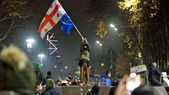 Ein Demonstrant steht mit georgischen National- und EU-Flaggen während einer Kundgebung vor dem Parlamentsgebäude in Tiflis, um gegen die Entscheidung der Regierung zu protestieren, die Verhandlungen über den Beitritt zur Europäischen Union für vier Jahre auszusetzen. © Zurab Tsertsvadze/AP/dpa 