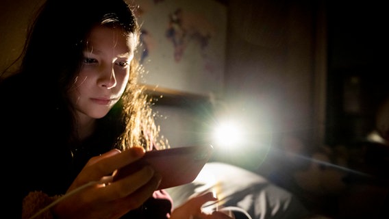 Ein Mädchen sitzt mit ihrem Handy auf dem Bett im dunkeln. © picture alliance / ROBIN UTRECHT | ROBIN UTRECHT 