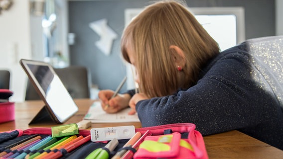 Ein Schuldkind sitzt im Januar 2021 im Elternhaus an einem Tisch im Homeschooling, auf der Federmappe im Vordergrund liegt eine Testkassette von einem negativen SARS-CoV-2-Schnelltest. © picture alliance / Fotostand | Fotostand / K. Schmitt 