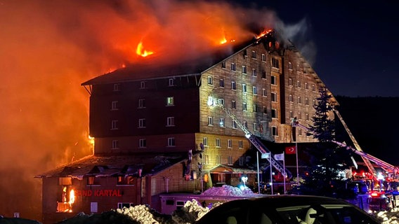 Flammen schlagen aus einem großen Hotelkomplex in einem türkischen Skigebiet. Davor sind Feuerwehrfahrzeuge mit Leitern zu sehen. © IHA/AP/dpa 