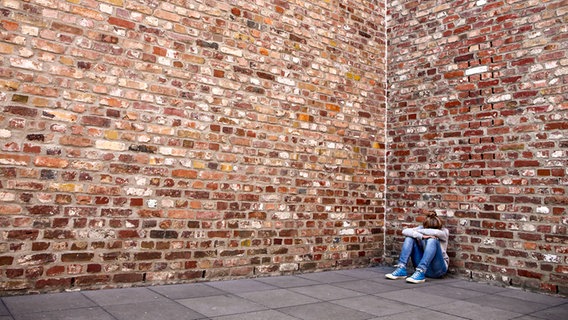 Ein junges Mädchen sitzt mit gesenktem Kopf vor einer hohen Mauer. © photocase.de Foto: Mr. Nico