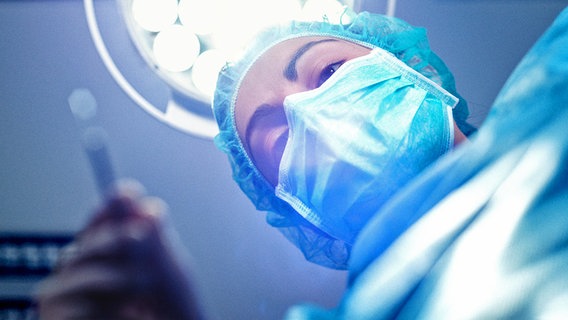 Aus Patientensicht nach oben: mit OP-Maske, Haube, Kittel und Handschuhen bekleidete Ärztin beugt sich mit einem Skalpell in der Hand nach unten. Dahinter eine OP-Lampe. © Addictive Stock / photocase.de 