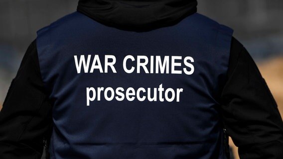 Ein Mann von hinten in einer dunklen Jacke mit der weißen Aufschrift auf dem Rücken "War Crimes prosecutor" © dpa/Zuma Press Foto: Carol Guzy