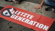 Aktivisten der "Letzten Generation" haben sich auf einer Straße festgeklebt, vor ihnen liegt ein Transparent mit dem Logo der Gruppe. © dpa-Bildfunk Foto: Swen Pförtner