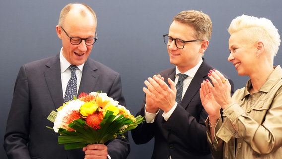 Friedrich Merz hält einen Blumenstrauß in der Hand, während Carsten Linnemann und Silvia Breher neben ihm stehen und klatschen. © dpa Foto: Michael Kappeler