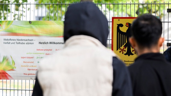 Zwei Männer stehen vor einem Banner mit Aufschrift „Herzlich Willkommen“ und einem Schild vom Bundesamt für Migration und Flüchtlinge die Landesaufnahmebehörde Niedersachsen am Standort Braunschweig. © dpa Foto: Julian Stratenschulte