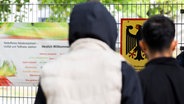 Zwei Männer stehen vor einem Banner mit Aufschrift „Herzlich Willkommen“ und einem Schild vom Bundesamt für Migration und Flüchtlinge die Landesaufnahmebehörde Niedersachsen am Standort Braunschweig. © dpa Foto: Julian Stratenschulte