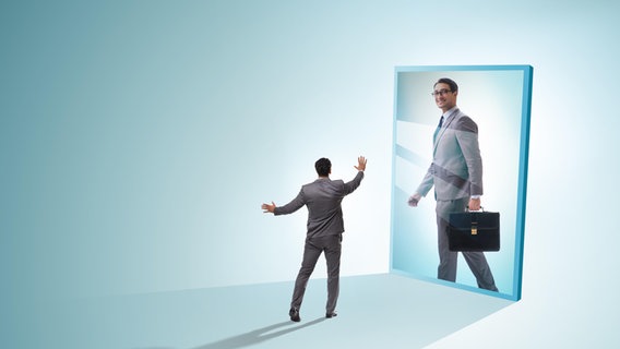 Ein Mann im Anzug betrachtet sich als erfolgreichen Geschäftsmann in einem vergrößerten Spiegel. © picture alliance / Zoonar | Elnur Amikishiyev 