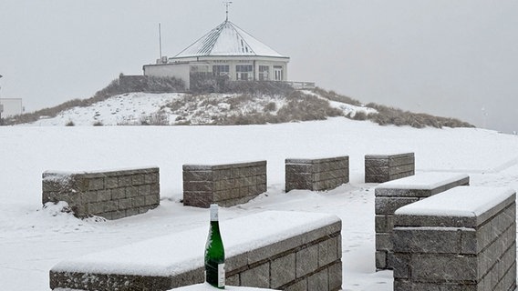 Eine Flasche steht auf einer mit Schnee bedeckten Bank vor dem Café Marienhöhe an der Hohen Düne auf der ostfriesischen Insel Norderney. Der Deutsche Wetterdienst erwartet heute für Ostfriesland und das Emsland leichten Schneefall - auch in den kommenden Tagen soll es in Niedersachsen mancherorts schneien. (Foto vom 12.2.2025) © Volker Bartels/dpa 