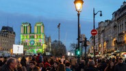 Fußgänger gehen in Paris an der angestrahlten Kathedrale Notre Dame vorbei. © Bernat Armangue/AP/dpa 