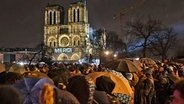 Mehrere Menschen stehen im Dunklen in der Nähe der Kathedrale Notre Dame in Paris, auf deren Fassade das Wort "Merci" projiziert wird. © dpa bildfunk/AP Foto: Bernat Armangue