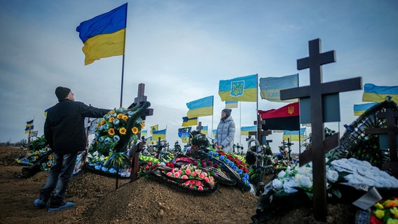 Anna, die Frau eines vor zwei Monaten getöteten Soldaten, und ihr Vater Oleksandr legen die ukrainische Nationalflagge am Grab ihres Mannes auf dem Friedhof der Hafenstadt Odessa nieder.  (Foto vom 24. Februar 2024) © Kay Nietfeld/dpa 