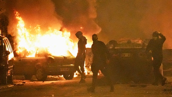 Demonstranten stehen in Paris auf einer Straße im Dunkeln, im Hintergrund sind brennende Fahrzeuge zu erkennen. © AP/dpa-Bildfunk Foto: Christophe Ena